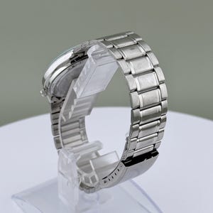Pu&ograve; includere: Un orologio da polso argentato con un cinturino in acciaio inossidabile. L'orologio &egrave; esposto su un supporto trasparente, evidenziando le maglie del cinturino e il quadrante. L'orologio ha un quadrante rotondo e una chiusura.
