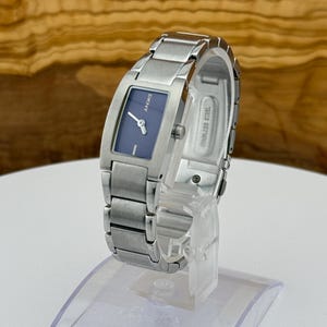 May include: A silver-toned DKNY watch with a rectangular navy blue face. The watch has a stainless steel band and is displayed on a clear stand. The watch face has silver hands and the DKNY logo.