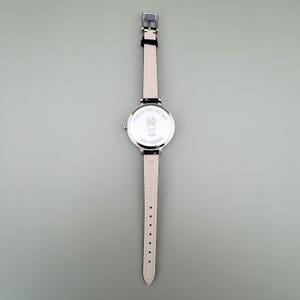 May include: A silver-toned wristwatch with a black leather strap and a light pink leather strap. The watch face features an owl design and the text "JAPAN MOVEMENT". The watch is on a gray background.