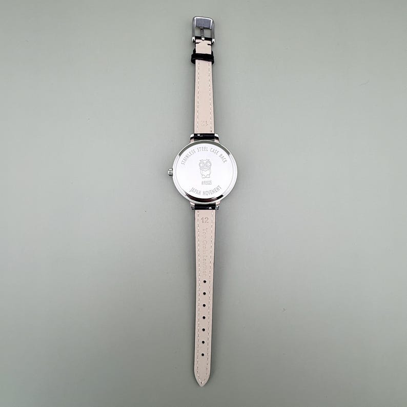 May include: A silver-toned wristwatch with a black leather strap and a light pink leather strap. The watch face features an owl design and the text "JAPAN MOVEMENT". The watch is on a gray background.