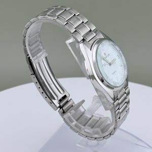 Pu&ograve; includere: Un orologio da polso in acciaio inossidabile argentato con quadrante bianco. L'orologio ha un movimento al quarzo e un'indicazione della data. Il cinturino &egrave; composto da maglie metalliche ad incastro. L'orologio &egrave; esposto su un supporto di plastica trasparente.