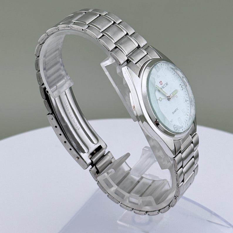 Pu&ograve; includere: Un orologio da polso in acciaio inossidabile argentato con quadrante bianco. L'orologio ha un movimento al quarzo e un'indicazione della data. Il cinturino &egrave; composto da maglie metalliche ad incastro. L'orologio &egrave; esposto su un supporto di plastica trasparente.