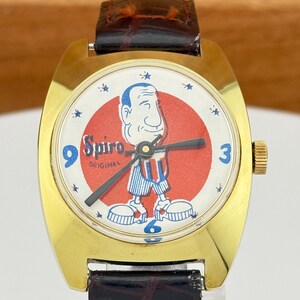 May include: A vintage gold-toned wristwatch with a brown leather band. The watch face features a cartoon character in red, white, and blue, with the word "Spiro" and the word "Original". The watch has blue numerals at the 3, 6, and 9 o'clock positions.