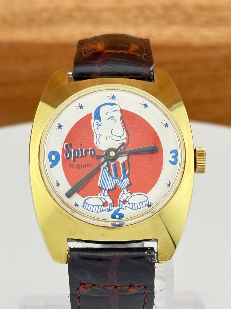 May include: A vintage gold-toned wristwatch with a brown leather band. The watch face features a cartoon character in red, white, and blue, with the word "Spiro" and the word "Original". The watch has blue numerals at the 3, 6, and 9 o'clock positions.