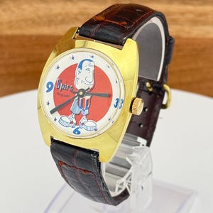 May include: Vintage gold-tone watch with a Spiro cartoon character on the face. The watch has a red and white dial, black hands, and a brown leather band. The watch is displayed on a clear stand.