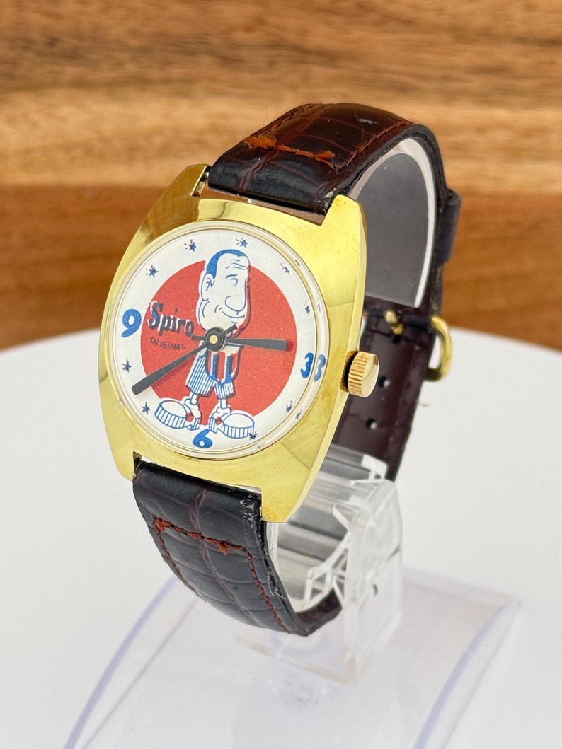 May include: Vintage gold-tone watch with a Spiro cartoon character on the face. The watch has a red and white dial, black hands, and a brown leather band. The watch is displayed on a clear stand.