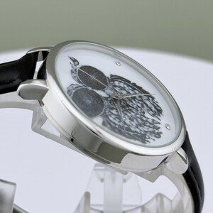 May include: Silver-tone wristwatch with a black leather band. The watch face features a black and white illustration of a bird and fruit. The watch has a round face with a crystal accent.