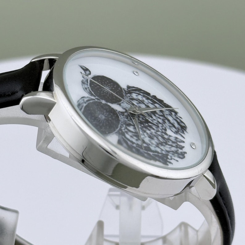 May include: Silver-tone wristwatch with a black leather band. The watch face features a black and white illustration of a bird and fruit. The watch has a round face with a crystal accent.