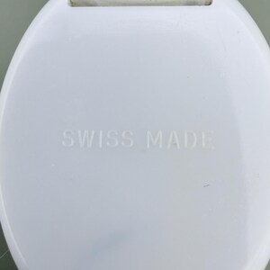 Puede incluir: Primer plano de un componente de reloj blanco y circular. El texto "SWISS MADE" est&aacute; grabado en la superficie. La parte superior del componente tiene una pieza de pl&aacute;stico blanco con el texto "15mm".