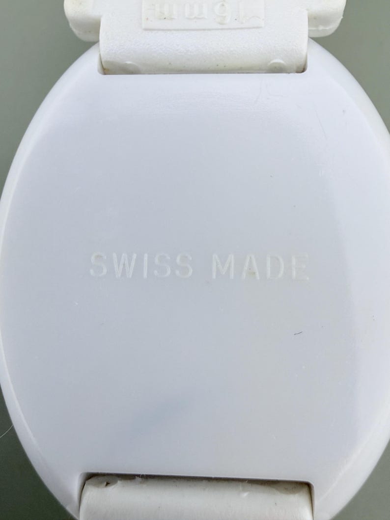 Puede incluir: Primer plano de un componente de reloj blanco y circular. El texto "SWISS MADE" est&aacute; grabado en la superficie. La parte superior del componente tiene una pieza de pl&aacute;stico blanco con el texto "15mm".