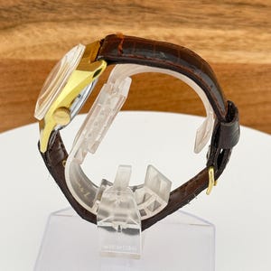 May include: A gold-toned wristwatch with a brown leather band. The watch face is visible, and the band is secured with a gold buckle. The watch is displayed on a clear plastic stand.