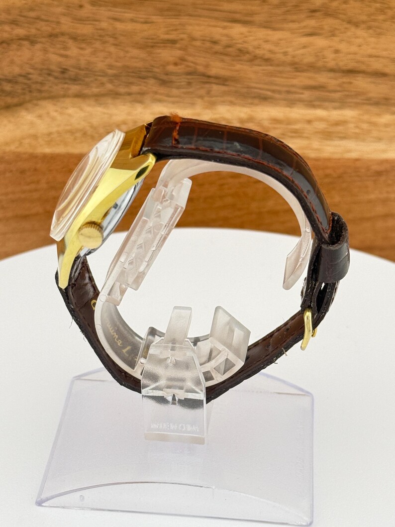 May include: A gold-toned wristwatch with a brown leather band. The watch face is visible, and the band is secured with a gold buckle. The watch is displayed on a clear plastic stand.