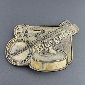 May include: A rectangular, brass-colored belt buckle featuring a banjo, violin, and guitar. The word "Bluegrass" is embossed on the buckle. The instruments are arranged on a textured background.