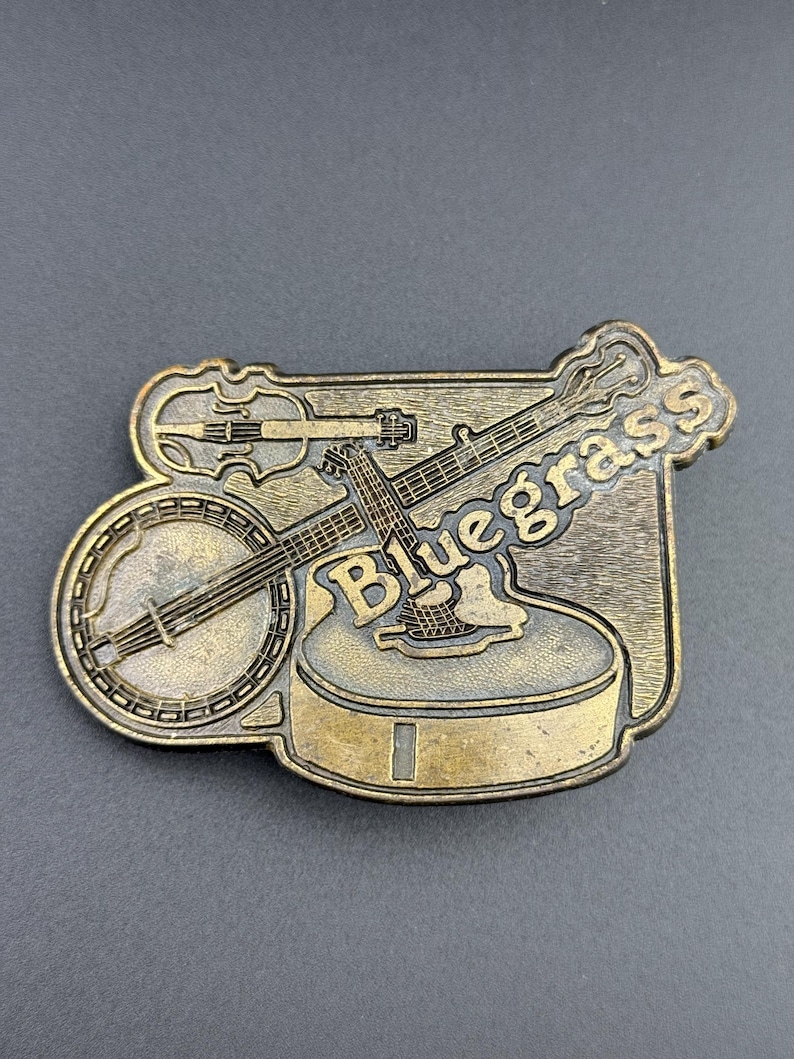 May include: A rectangular, brass-colored belt buckle featuring a banjo, violin, and guitar. The word "Bluegrass" is embossed on the buckle. The instruments are arranged on a textured background.