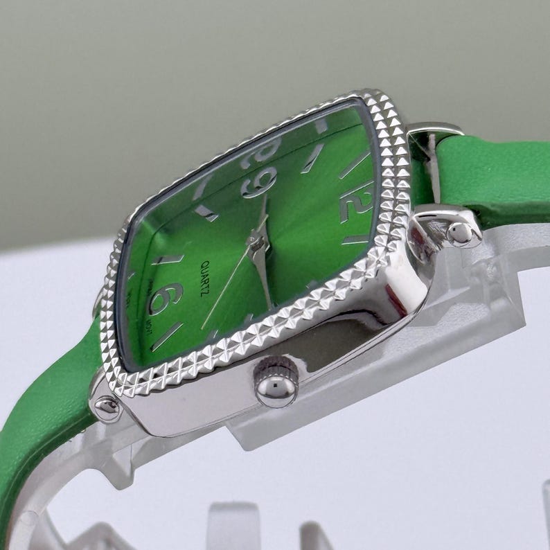 May include: A green quartz watch with a silver bezel and a green leather strap. The watch face is a bright green with silver hands and numerals. The watch has a rectangular face with a decorative edge.