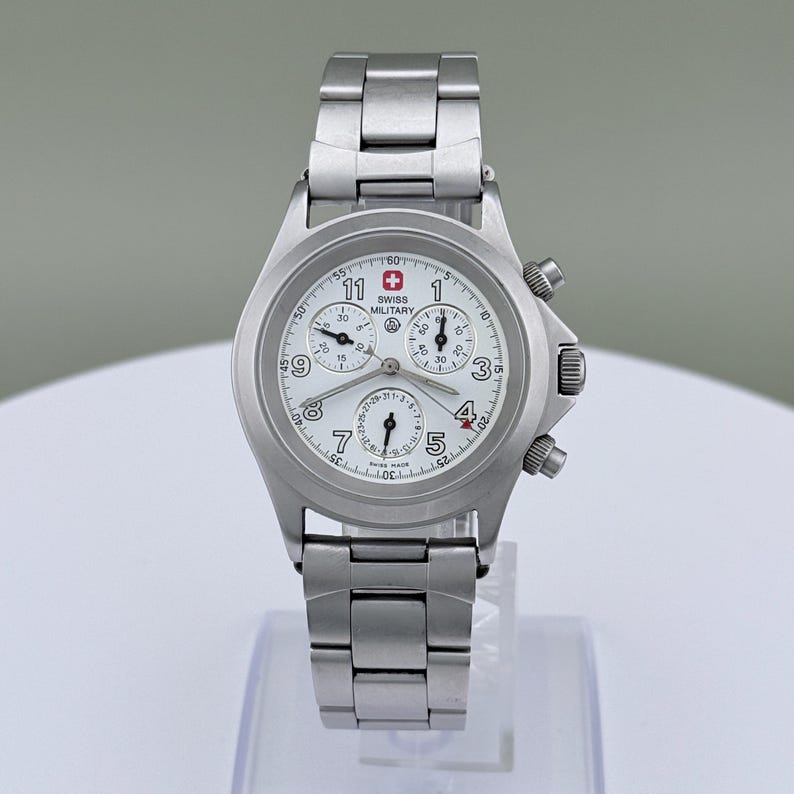 Puede incluir: Un reloj Swiss Military de acero inoxidable con esfera blanca. El reloj cuenta con un cron&oacute;grafo con m&uacute;ltiples subesferas, una pantalla de fecha y agujas luminosas. La correa y la caja son de acero inoxidable.
