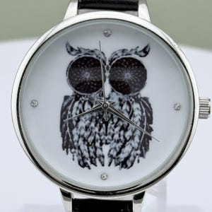 May include: Silver-tone wristwatch with a black leather band. The watch face features a black and white owl design with three small crystal accents. The watch hands are silver, and the watch has a silver bezel.