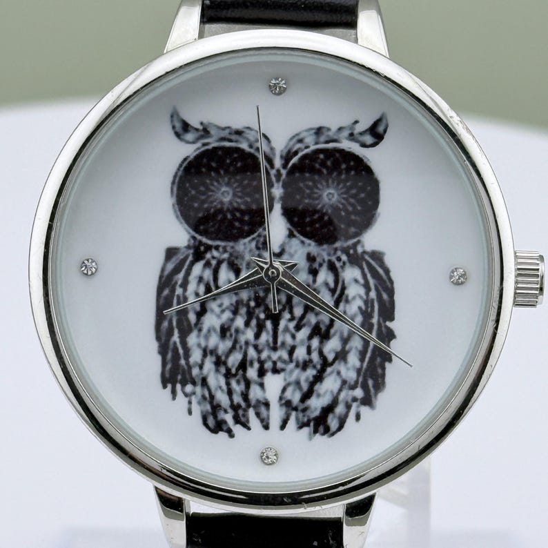 May include: Silver-tone wristwatch with a black leather band. The watch face features a black and white owl design with three small crystal accents. The watch hands are silver, and the watch has a silver bezel.