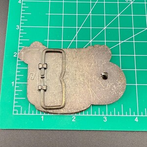 May include: A metal belt buckle with a rectangular frame and a rounded, irregular shape. The buckle is a dull silver color with a dark, aged appearance. The buckle is approximately 3 inches long.