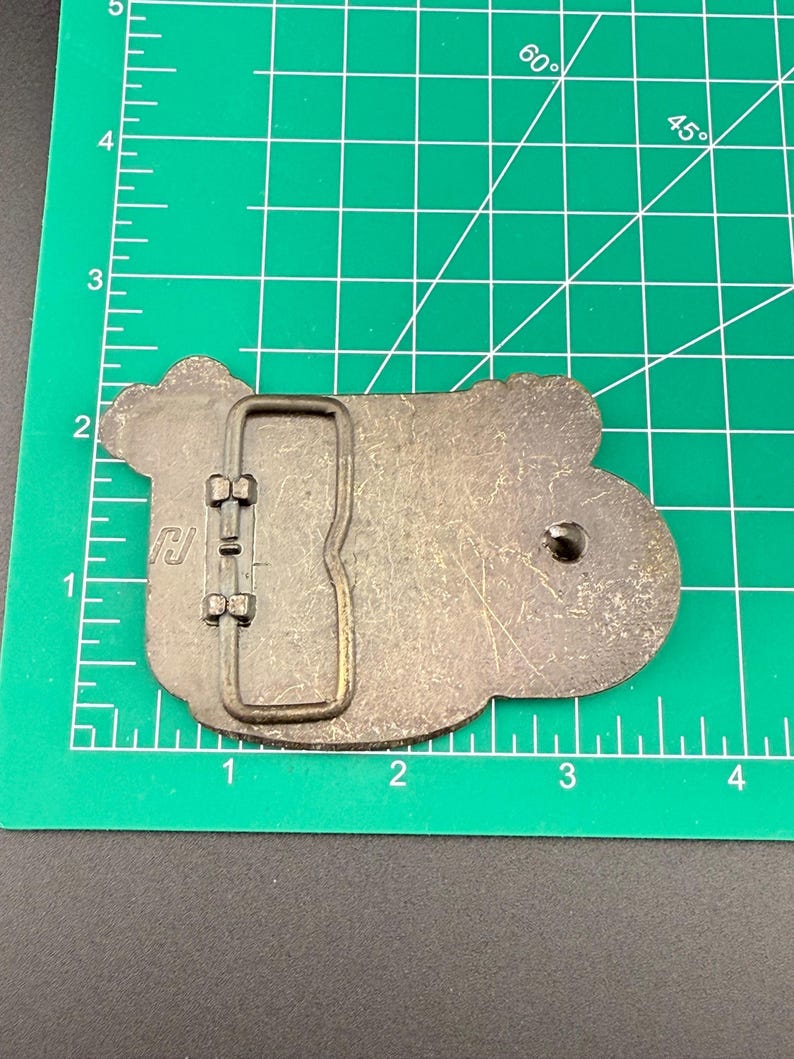 May include: A metal belt buckle with a rectangular frame and a rounded, irregular shape. The buckle is a dull silver color with a dark, aged appearance. The buckle is approximately 3 inches long.