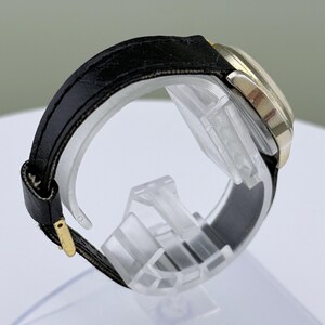 K&ouml;nnte beinhalten: Seitenansicht einer goldfarbenen Armbanduhr mit schwarzem Lederarmband. Das Zifferblatt der Uhr ist rund, und das Armband wird mit einer goldfarbenen Schnalle befestigt. Die Uhr wird auf einem transparenten Kunststoffst&auml;nder pr&auml;sentiert.