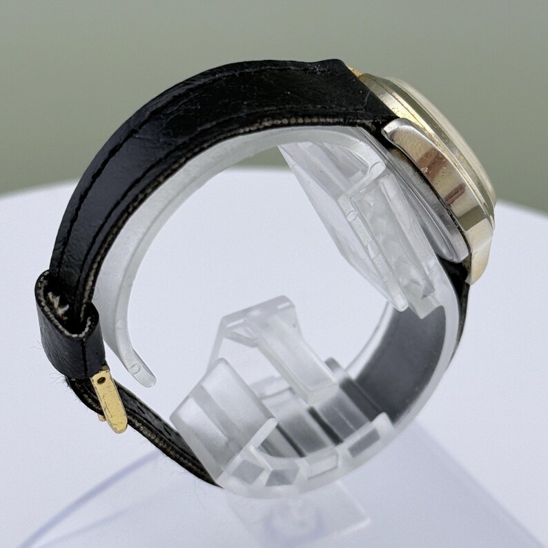 K&ouml;nnte beinhalten: Seitenansicht einer goldfarbenen Armbanduhr mit schwarzem Lederarmband. Das Zifferblatt der Uhr ist rund, und das Armband wird mit einer goldfarbenen Schnalle befestigt. Die Uhr wird auf einem transparenten Kunststoffst&auml;nder pr&auml;sentiert.