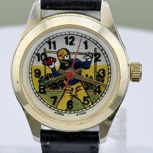 K&ouml;nnte beinhalten: Vintage Armbanduhr mit goldfarbenem Geh&auml;use und schwarzem Lederarmband. Das Zifferblatt zeigt eine Footballspieler-Grafik in Gelb, Blau und Rot. Das Zifferblatt hat schwarze Zahlen und die Worte "HONG KONG" unten.