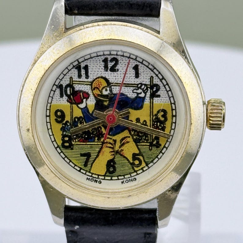 K&ouml;nnte beinhalten: Vintage Armbanduhr mit goldfarbenem Geh&auml;use und schwarzem Lederarmband. Das Zifferblatt zeigt eine Footballspieler-Grafik in Gelb, Blau und Rot. Das Zifferblatt hat schwarze Zahlen und die Worte "HONG KONG" unten.