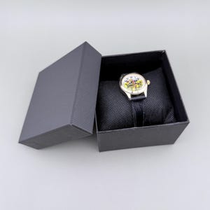 K&ouml;nnte beinhalten: Eine Armbanduhr mit farbenfrohem Zifferblatt und schwarzem Lederarmband liegt auf einem schwarzen Kissen in einer dunkelgrauen Geschenkbox. Das Zifferblatt der Uhr hat ein Design mit gelben, roten und gr&uuml;nen Akzenten. Die Box ist ge&ouml;ffnet und zeigt die Uhr.
