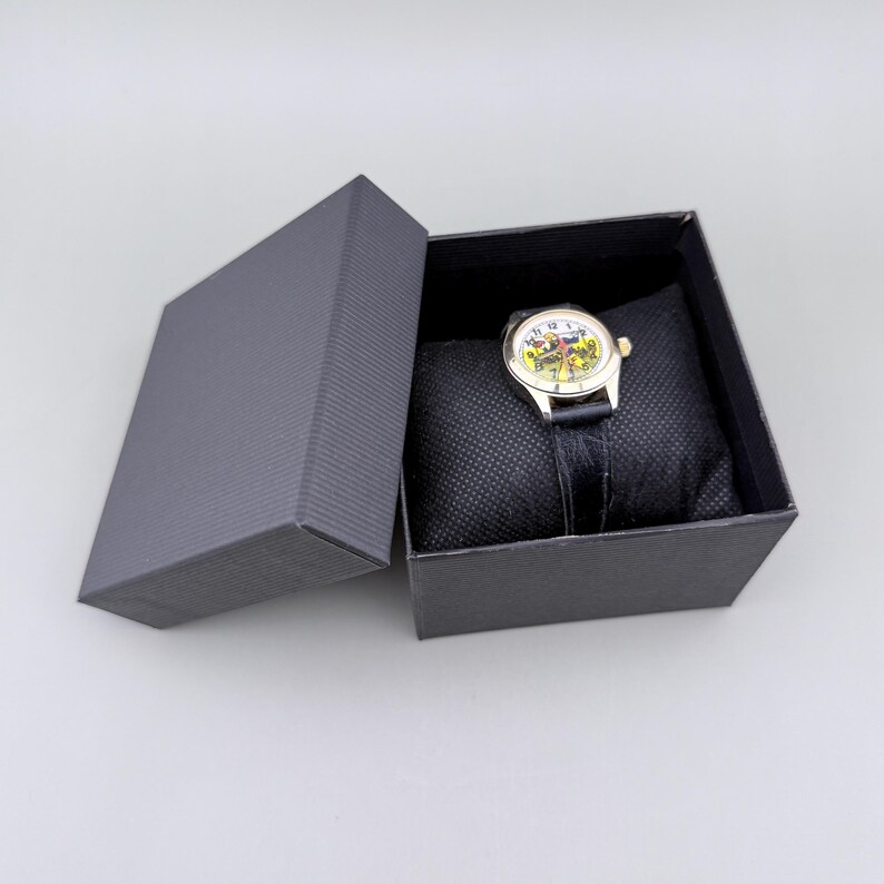 K&ouml;nnte beinhalten: Eine Armbanduhr mit farbenfrohem Zifferblatt und schwarzem Lederarmband liegt auf einem schwarzen Kissen in einer dunkelgrauen Geschenkbox. Das Zifferblatt der Uhr hat ein Design mit gelben, roten und gr&uuml;nen Akzenten. Die Box ist ge&ouml;ffnet und zeigt die Uhr.