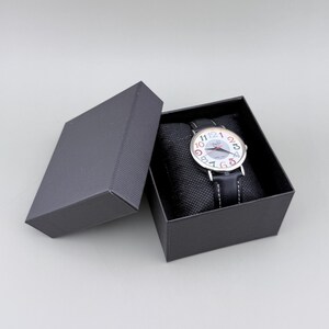 May include: A wristwatch with a white face and colorful numerals is displayed in an open black box. The watch has a black leather band and is presented on a black cushion. The box is on a gray surface.