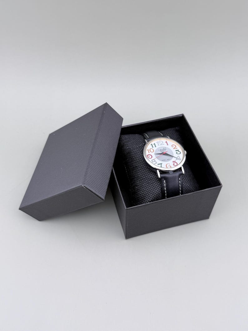 May include: A wristwatch with a white face and colorful numerals is displayed in an open black box. The watch has a black leather band and is presented on a black cushion. The box is on a gray surface.