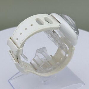 Puede incluir: Un reloj de pulsera blanco con una esfera transparente y redondeada y una correa blanca. La correa tiene m&uacute;ltiples agujeros para ajustar y un cierre de hebilla. El reloj se exhibe sobre un soporte de pl&aacute;stico transparente.