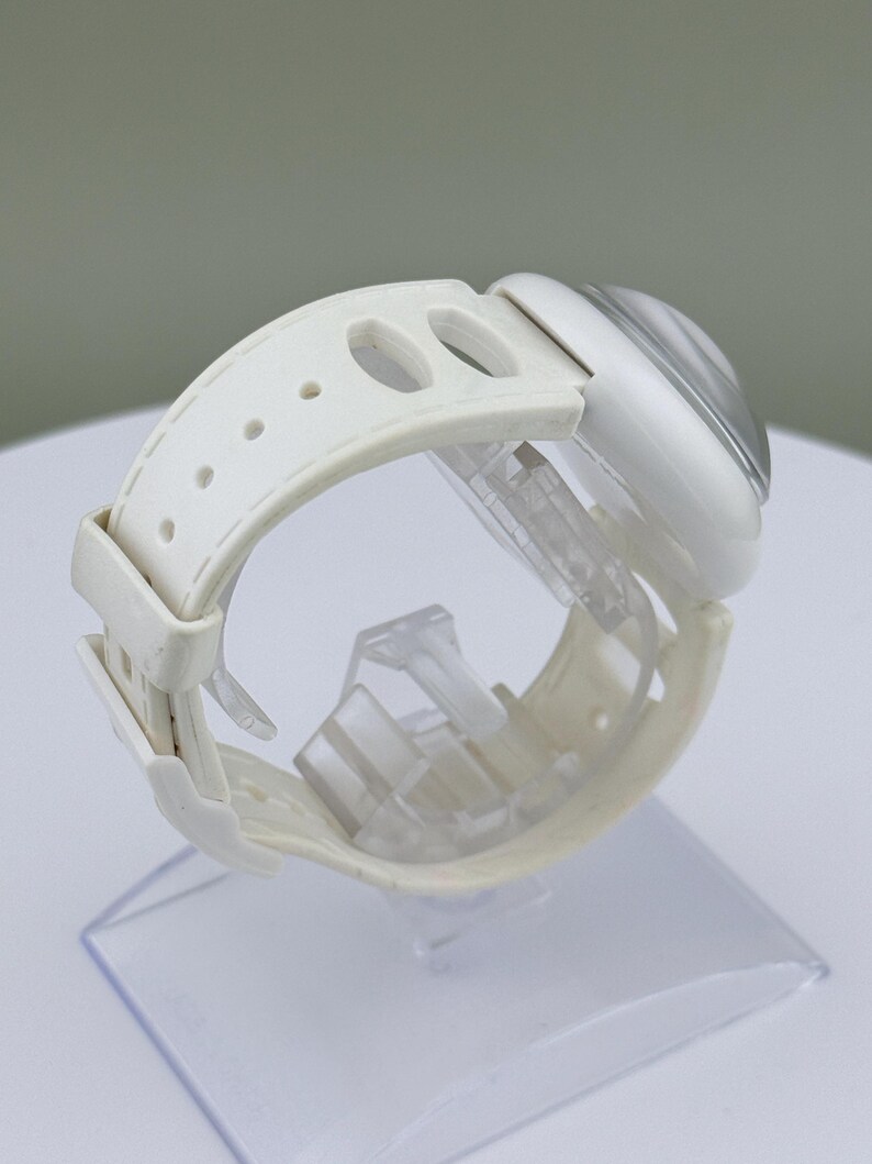 Puede incluir: Un reloj de pulsera blanco con una esfera transparente y redondeada y una correa blanca. La correa tiene m&uacute;ltiples agujeros para ajustar y un cierre de hebilla. El reloj se exhibe sobre un soporte de pl&aacute;stico transparente.