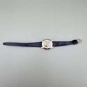 K&ouml;nnte beinhalten: Eine goldfarbene Armbanduhr mit einem schwarzen Lederarmband. Das Zifferblatt der Uhr ist wei&szlig; mit schwarzen Ziffern und Zeigern. Das Armband wird mit einer goldfarbenen Schnalle befestigt. Die Uhr liegt auf einer grauen Oberfl&auml;che.