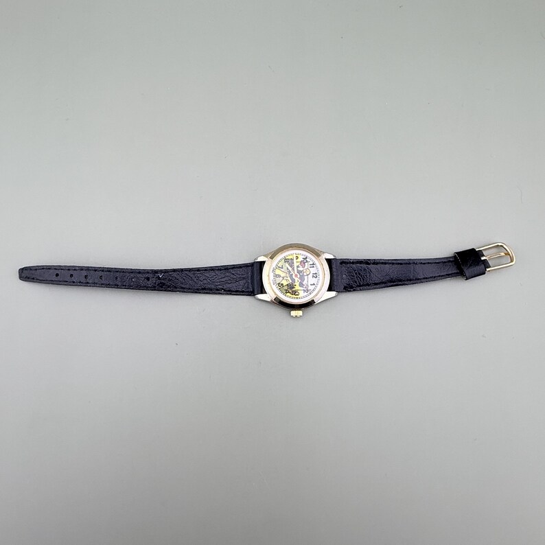 K&ouml;nnte beinhalten: Eine goldfarbene Armbanduhr mit einem schwarzen Lederarmband. Das Zifferblatt der Uhr ist wei&szlig; mit schwarzen Ziffern und Zeigern. Das Armband wird mit einer goldfarbenen Schnalle befestigt. Die Uhr liegt auf einer grauen Oberfl&auml;che.