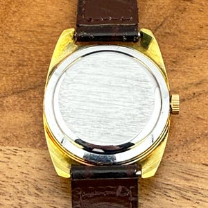 May include: A vintage gold-toned wristwatch with a brown leather strap. The watch face is silver, and the back is visible, showing the watch's inner workings. The strap is marked "Genuine". The watch is set against a wooden surface.