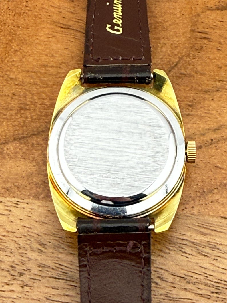 May include: A vintage gold-toned wristwatch with a brown leather strap. The watch face is silver, and the back is visible, showing the watch's inner workings. The strap is marked "Genuine". The watch is set against a wooden surface.