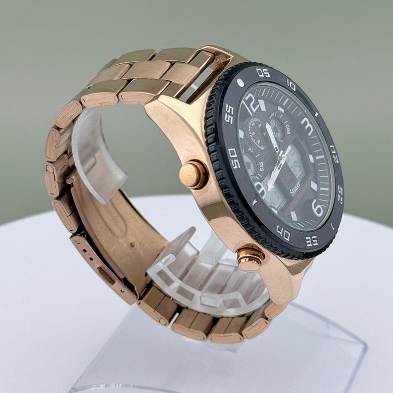K&ouml;nnte beinhalten: Eine ros&eacute;goldfarbene Armbanduhr mit schwarzem Zifferblatt und passendem Gliederarmband. Das Zifferblatt zeigt mehrere Hilfszifferbl&auml;tter und digitale Anzeigen. Die Uhr wird auf einem transparenten Kunststoffst&auml;nder pr&auml;sentiert.
