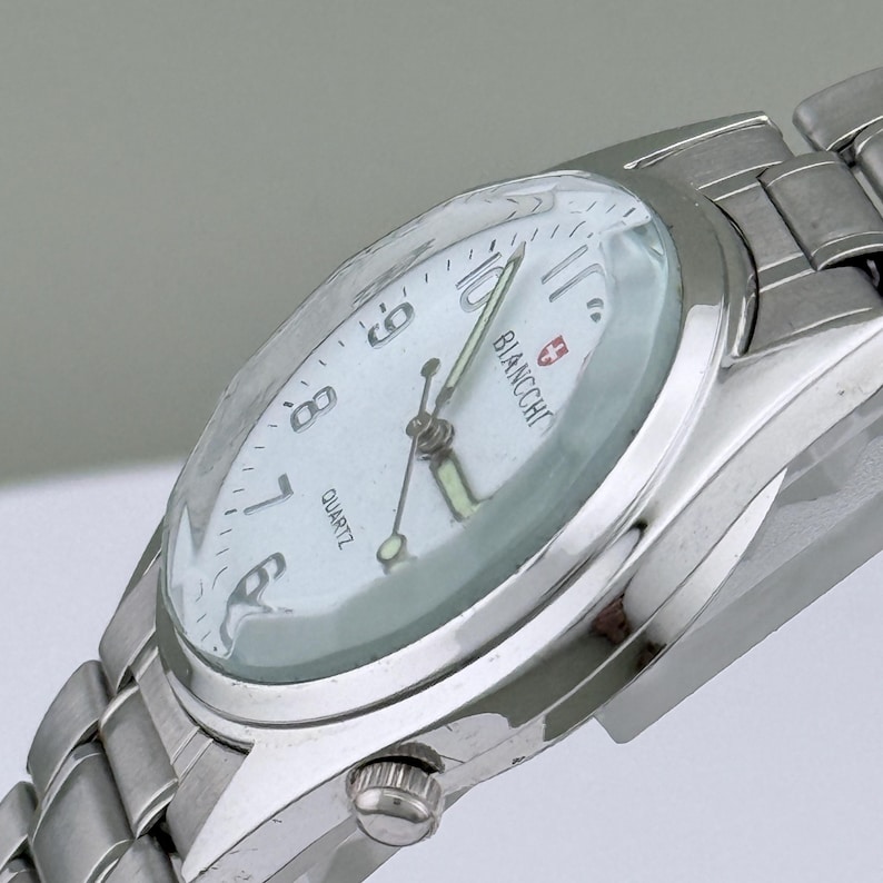 Pu&ograve; includere: Un orologio da polso argentato con quadrante bianco. Il quadrante mostra il marchio "BIANCHCHI" e la parola "QUARTZ". L'orologio ha un cinturino argentato e una piccola corona per regolare l'ora.