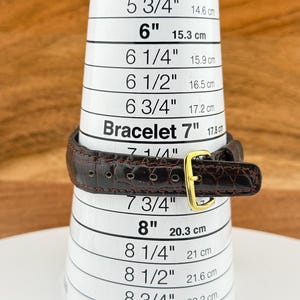 May include: A brown leather bracelet with a gold-colored buckle is measured on a white EZ Bracelet Mini sizing tool. The tool displays measurements in inches and centimeters, with the bracelet fitting at approximately 7 inches (17.8 cm).
