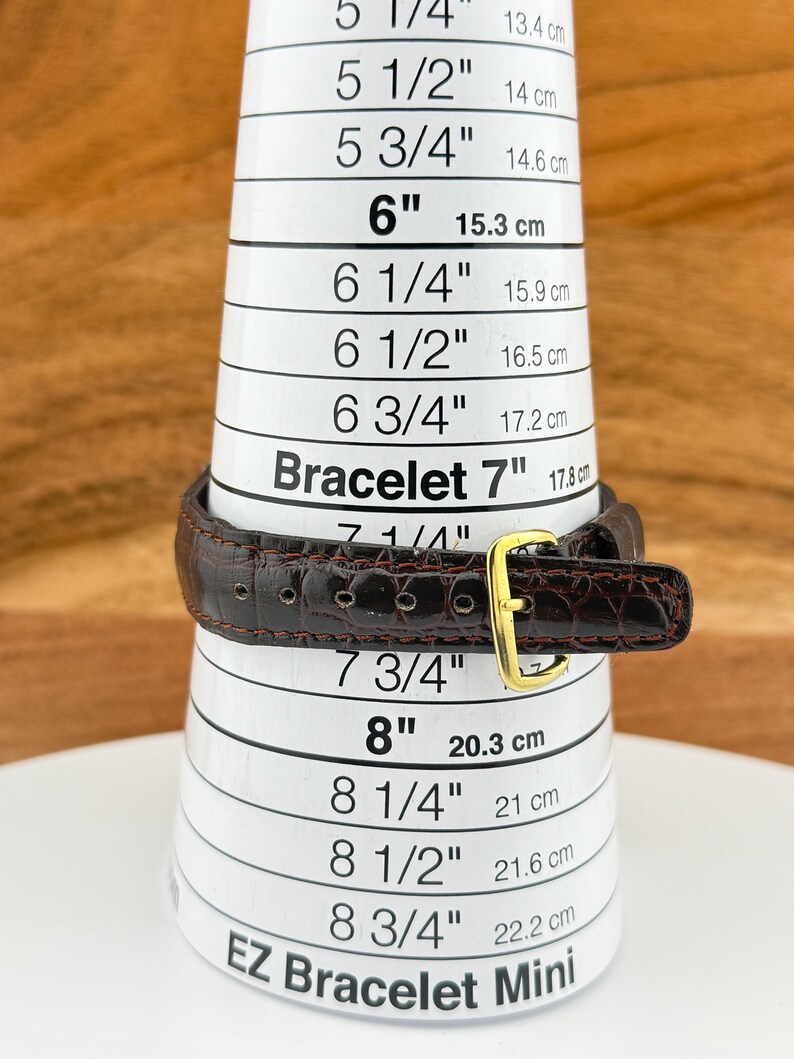 May include: A brown leather bracelet with a gold-colored buckle is measured on a white EZ Bracelet Mini sizing tool. The tool displays measurements in inches and centimeters, with the bracelet fitting at approximately 7 inches (17.8 cm).