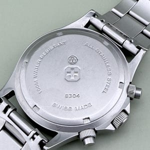 Puede incluir: Primer plano de la parte trasera de un reloj de pulsera de acero inoxidable. La esfera del reloj es plateada con el texto "100M WATER RESISTANT" y "ALL STAINLESS STEEL". El reloj tambi&eacute;n tiene el texto "SWISS MADE" y el n&uacute;mero "8304".