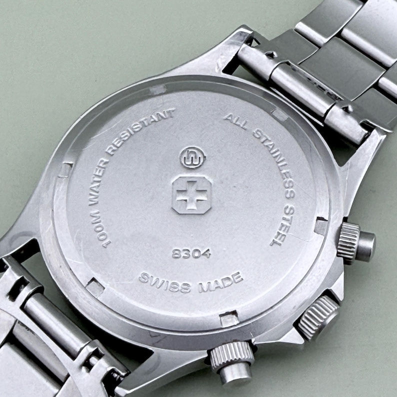 Puede incluir: Primer plano de la parte trasera de un reloj de pulsera de acero inoxidable. La esfera del reloj es plateada con el texto "100M WATER RESISTANT" y "ALL STAINLESS STEEL". El reloj tambi&eacute;n tiene el texto "SWISS MADE" y el n&uacute;mero "8304".