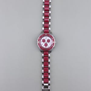 May include: A stainless steel watch with a red bezel and matching red links on the band. The watch face is white with red accents and three sub-dials. The watch band is approximately 7 inches long.