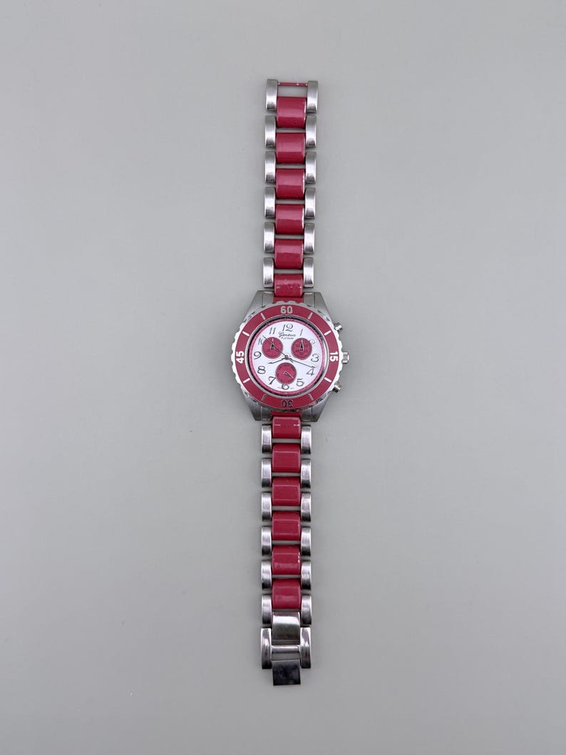 May include: A stainless steel watch with a red bezel and matching red links on the band. The watch face is white with red accents and three sub-dials. The watch band is approximately 7 inches long.