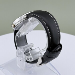May include: A silver-toned wristwatch with a black leather band and white stitching. The watch is displayed on a clear plastic stand, showcasing the band's texture and the watch's face. The watch has a classic design.