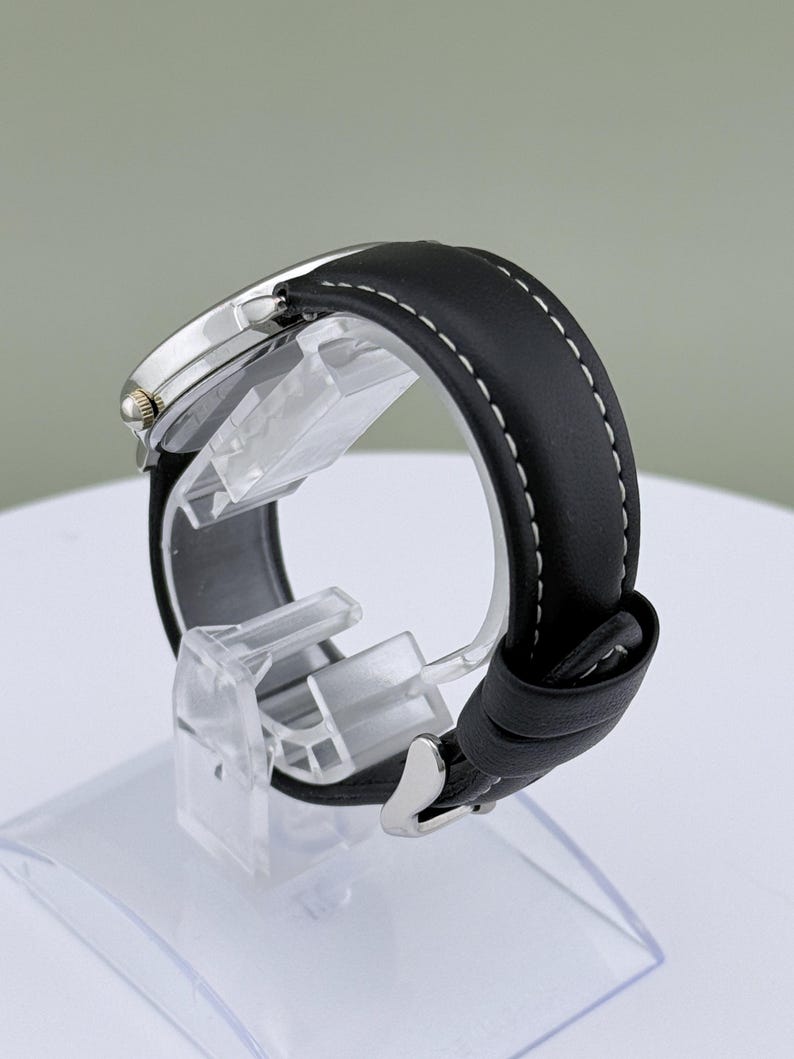 May include: A silver-toned wristwatch with a black leather band and white stitching. The watch is displayed on a clear plastic stand, showcasing the band's texture and the watch's face. The watch has a classic design.