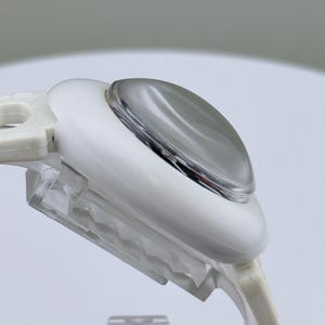 Puede incluir: Primer plano de un dispositivo de pl&aacute;stico blanco y transparente con una lente redondeada de color plateado. El dispositivo tiene una forma curva y est&aacute; dise&ntilde;ado con una base transparente y texturizada. El fondo es gris neutro suave.
