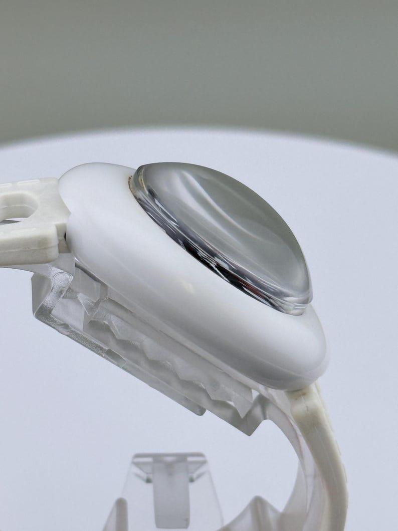 Puede incluir: Primer plano de un dispositivo de pl&aacute;stico blanco y transparente con una lente redondeada de color plateado. El dispositivo tiene una forma curva y est&aacute; dise&ntilde;ado con una base transparente y texturizada. El fondo es gris neutro suave.